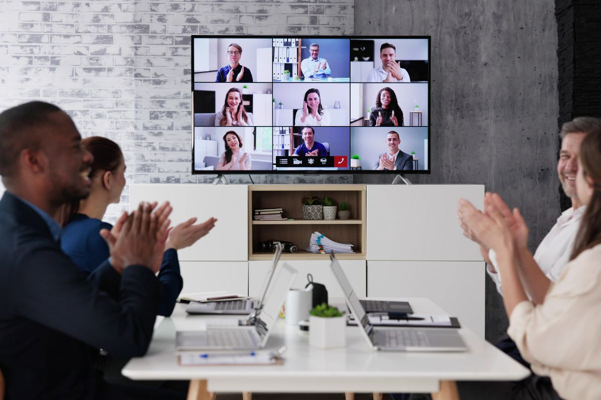 People in an office clapping during a hybrid meeting, with participants visible on a screen via video conference.